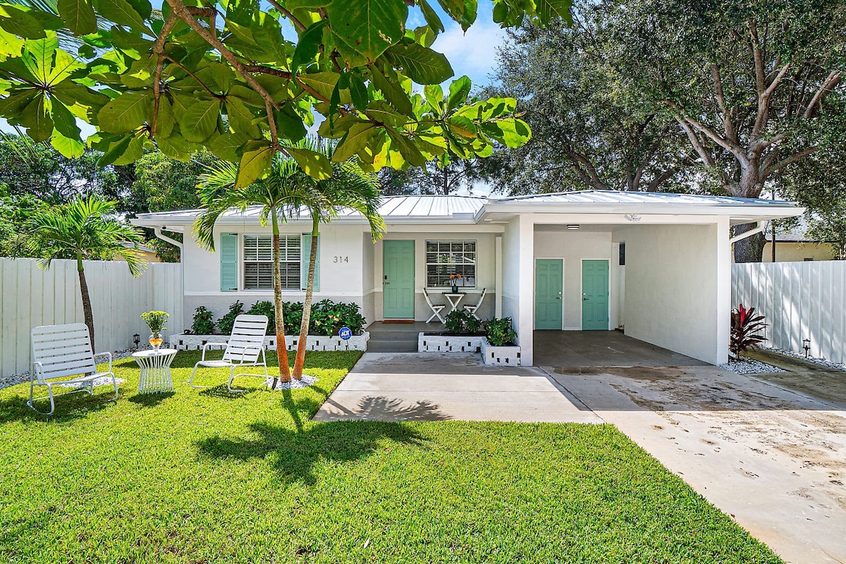 A charming casita is set against a lush green lawn, framed by vibrant foliage. The entrance features light blue accents and a small seating area with two white chairs, inviting relaxation. Sunlight casts gentle shadows, enhancing the welcoming appearance of the home's exterior.