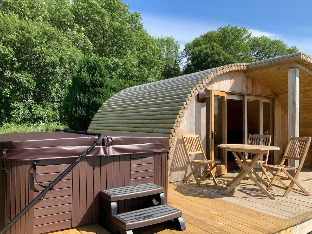 A private hot tub is positioned on a wooden deck beside a charming glamping coop. A round table and two chairs are arranged nearby, surrounded by lush greenery. The coop features a curved wood exterior and glass doors, providing a connection to the tranquil outdoor setting.