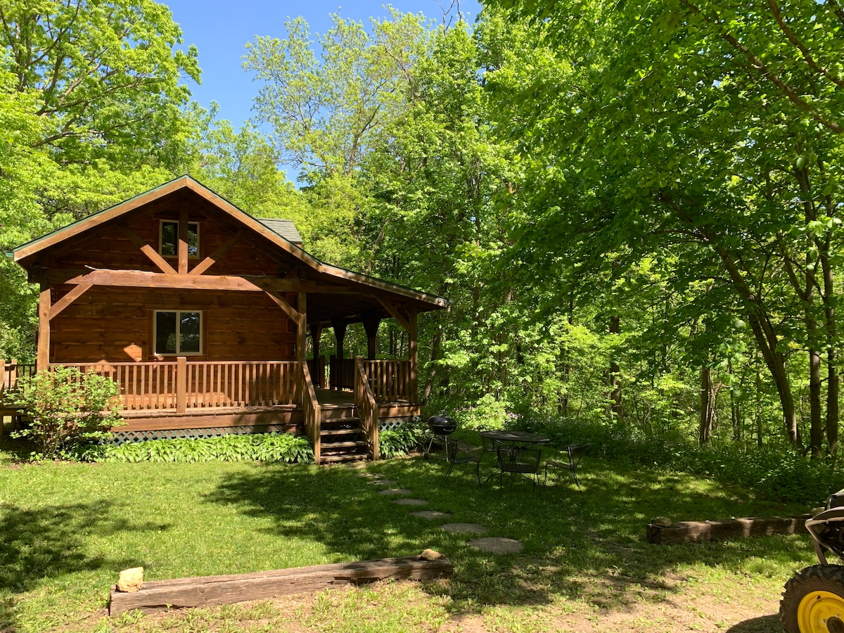 A log cabin is surrounded by dense greenery in a wooded area, featuring a welcoming front porch with a railing. The grassy yard includes a seating area and a fire pit, blending harmoniously with the natural landscape.