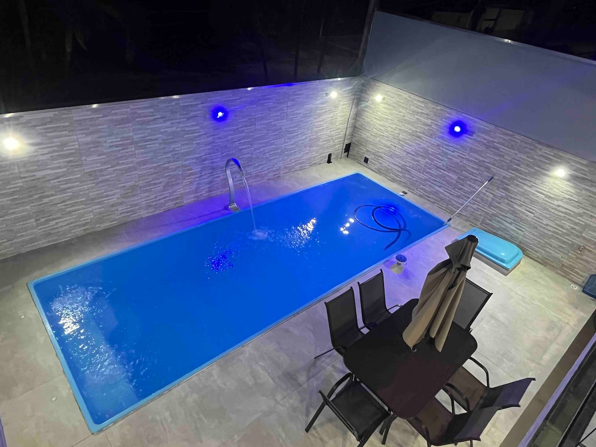 An outdoor area features a rectangular swimming pool illuminated with blue lights. A set of dark chairs and a table is positioned nearby, complemented by an umbrella. The pool area is framed by modern stone walls and is well-lit, enhancing visibility.