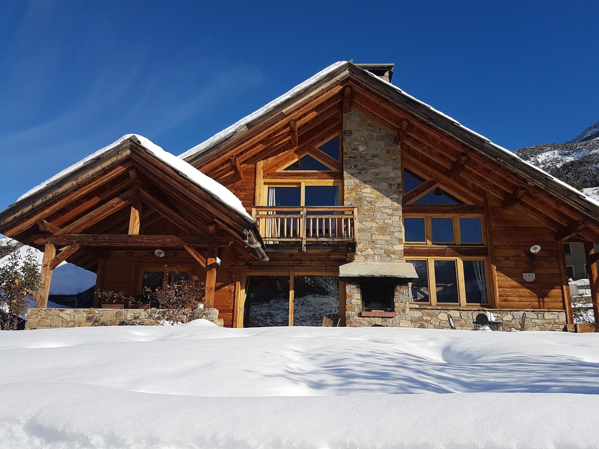 The chalet features a striking combination of natural stone and wood, with a sloped roof and expansive windows that invite natural light. A covered terrace provides outdoor seating, while fresh snow blankets the ground, creating a serene winter scene.