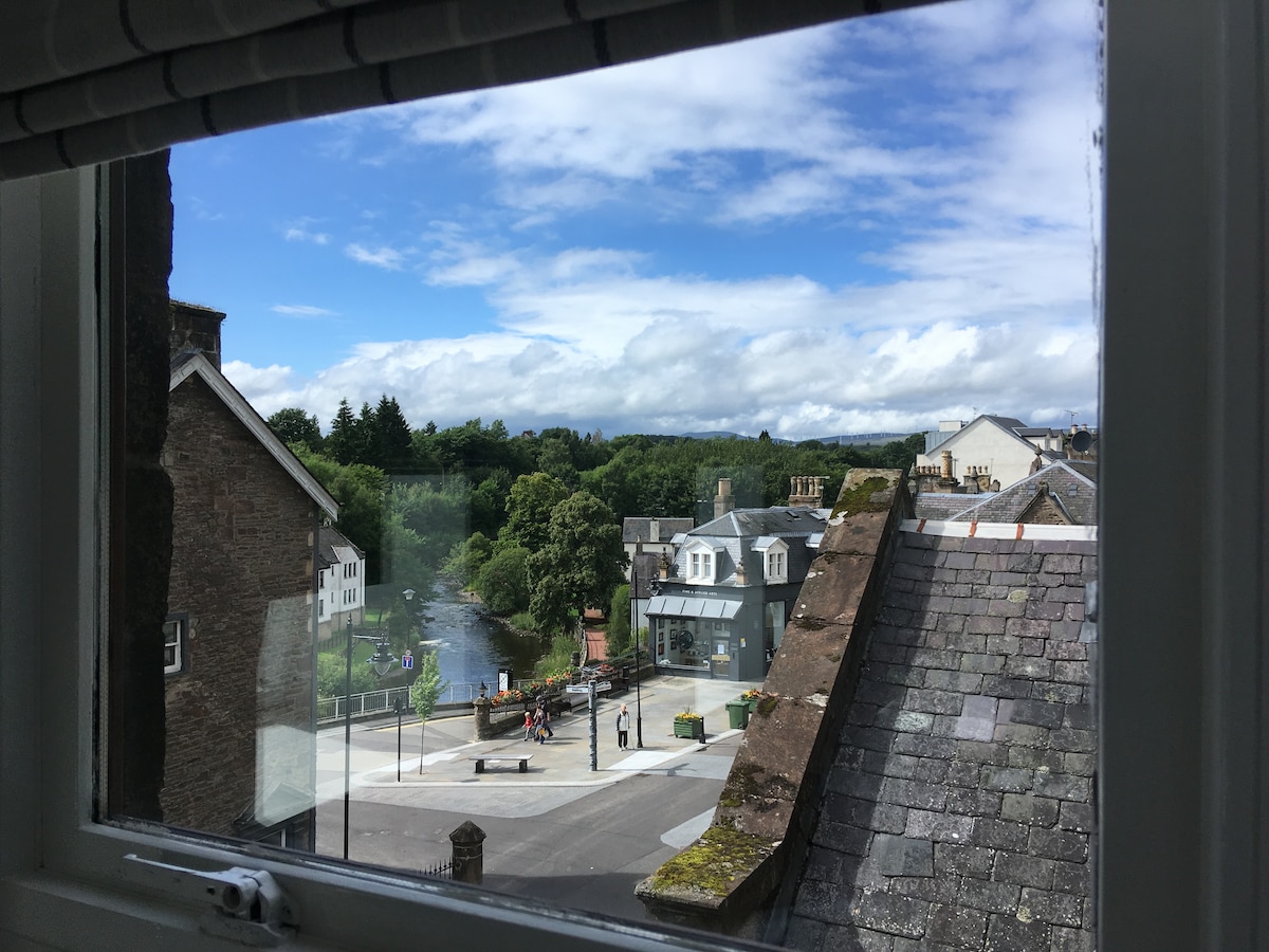 A view from a window reveals a vibrant street scene with lush greenery and the River Allan in the background. The surrounding buildings display diverse architectural styles, while the sky is filled with clouds and patches of blue, enhancing the scenic outlook.
