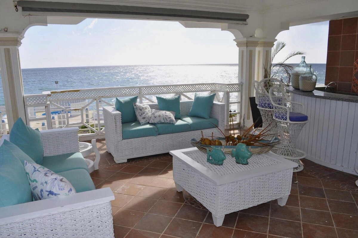 An open terrace features white wicker furniture, including a spacious seating area with soft blue cushions. A white coffee table with decorative items sits between the seating. A bar area with high stools overlooks the sea, offering a serene view of the water.