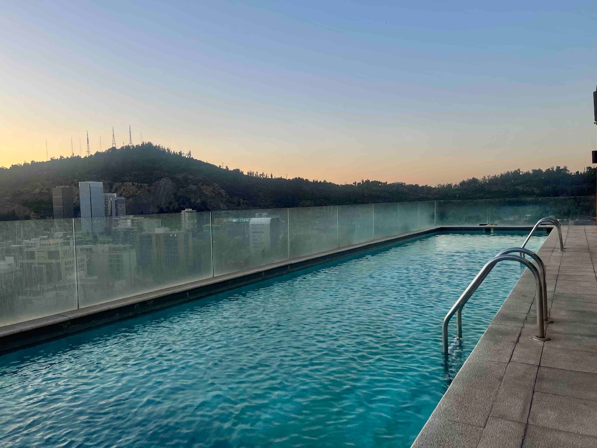 An infinity pool is situated on a rooftop, reflecting the tranquil blue water. Surrounding glass panels provide unobstructed views of a distant hillside, with soft hues of sunset lightening the sky.