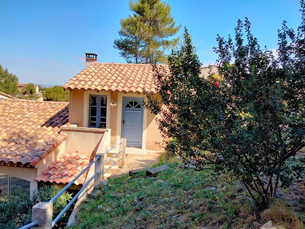 A charming studio is nestled within a hillside setting, featuring a terracotta-tiled roof and light-colored exterior. The entrance showcases a classic arched door framed by two windows. Surrounding greenery includes shrubs and trees, contributing to the serene residential atmosphere.