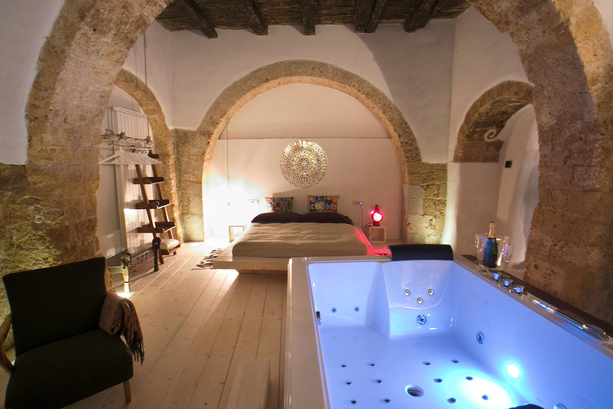 A spacious room features a wooden ceiling with exposed beams and stone arches. A two-person jacuzzi with ambient lighting is positioned in the foreground. A large bed is situated at the center of the space, complemented by decorative wall art and soft lighting.