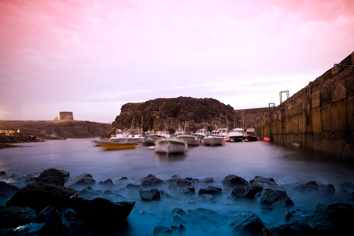 A serene harbor scene is captured during twilight, showcasing several boats gently bobbing on calm waters. Rocky formations line the shoreline, while a historical structure is visible in the background, surrounded by a tranquil atmosphere.