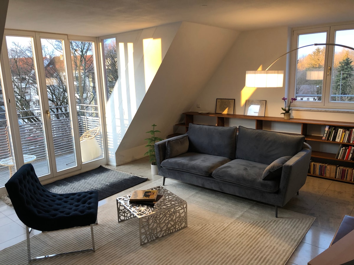 A bright living area features a modern gray sofa and a unique chair with a textured surface. Light is streamed in through large windows, reflecting off a decorative metal coffee table. A bookshelf filled with various books and a small plant are visible in the background.
