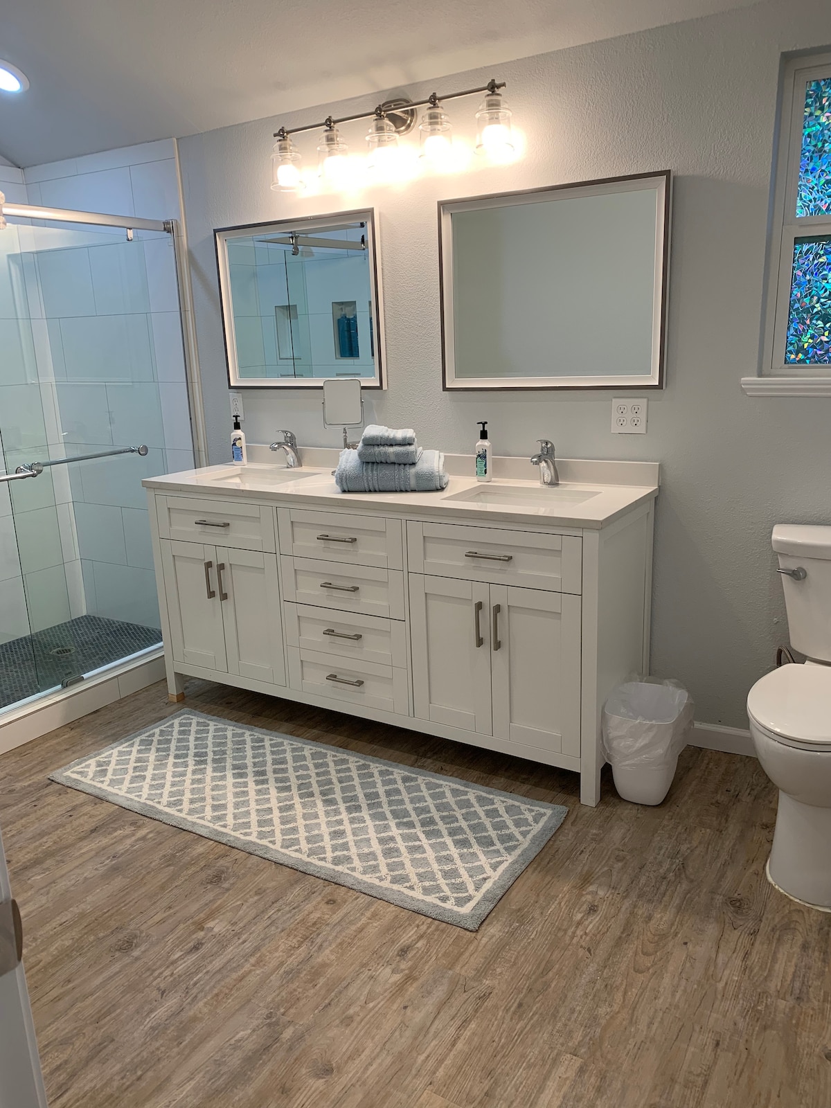 A spacious bathroom features dual sinks with modern fixtures and stylish cabinetry. Natural light filters through a nearby window. A glass-enclosed shower is visible to the left, alongside a light-colored area rug placed on a wood-style floor.