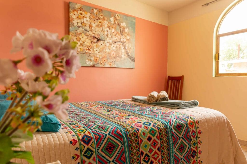 A warm-toned bedroom features a spacious bed adorned with a colorful patterned blanket. Fresh towels are neatly placed at the foot of the bed. A floral artwork adds a touch of charm above the bed, while a soft light filters through an arched window.