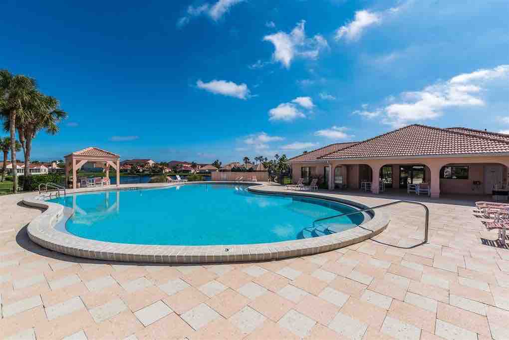The outdoor pool area is surrounded by palm trees and features a spacious deck of light-colored pavers. The pool has a curved shape and is complemented by a nearby clubhouse with a terracotta roof, offering shaded seating options.