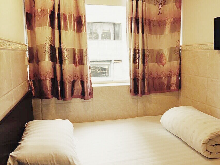 A bright sleeping area is highlighted by large windows draped with patterned curtains. A spacious bed is positioned against a wall, featuring two neatly arranged pillows. Natural light creates a serene environment, enhancing the simplicity of the decor.