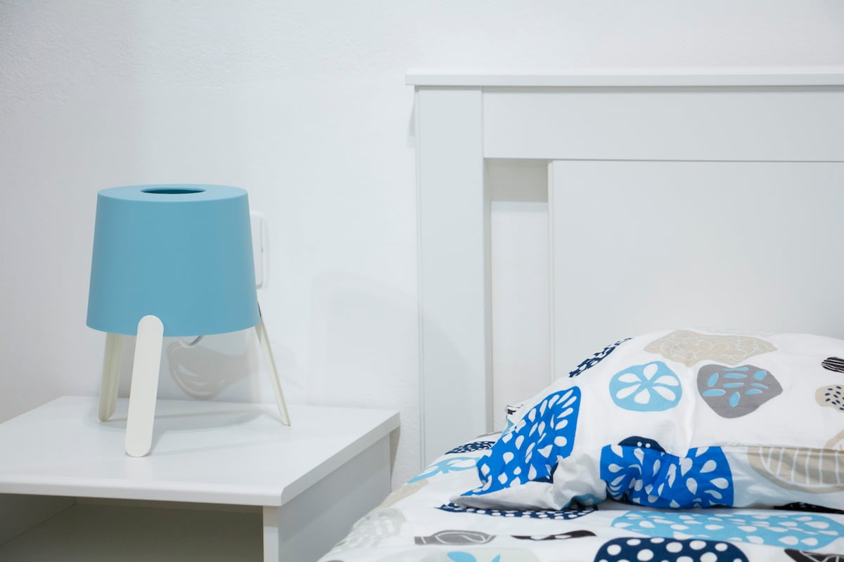 A bedside table is positioned next to a bed adorned with a patterned duvet cover featuring various shapes in blue and neutral tones. A modern blue lamp with three legs sits atop the table, providing a functional accent.