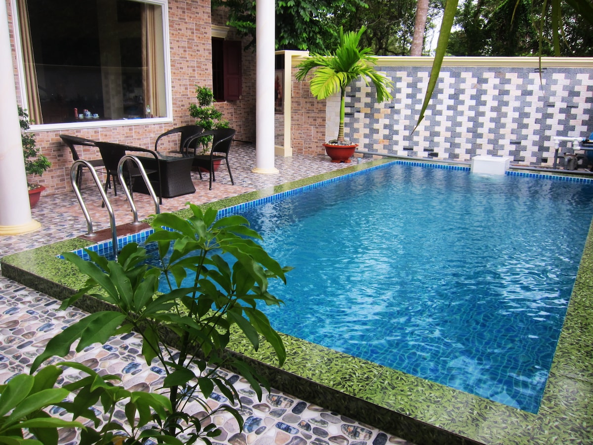 A private swimming pool is surrounded by lush greenery, featuring a tiled border. Comfortable outdoor seating is positioned nearby, enhancing the space's inviting feel. The pool's clear blue water reflects the surrounding plants and walls, providing a serene atmosphere for relaxation.