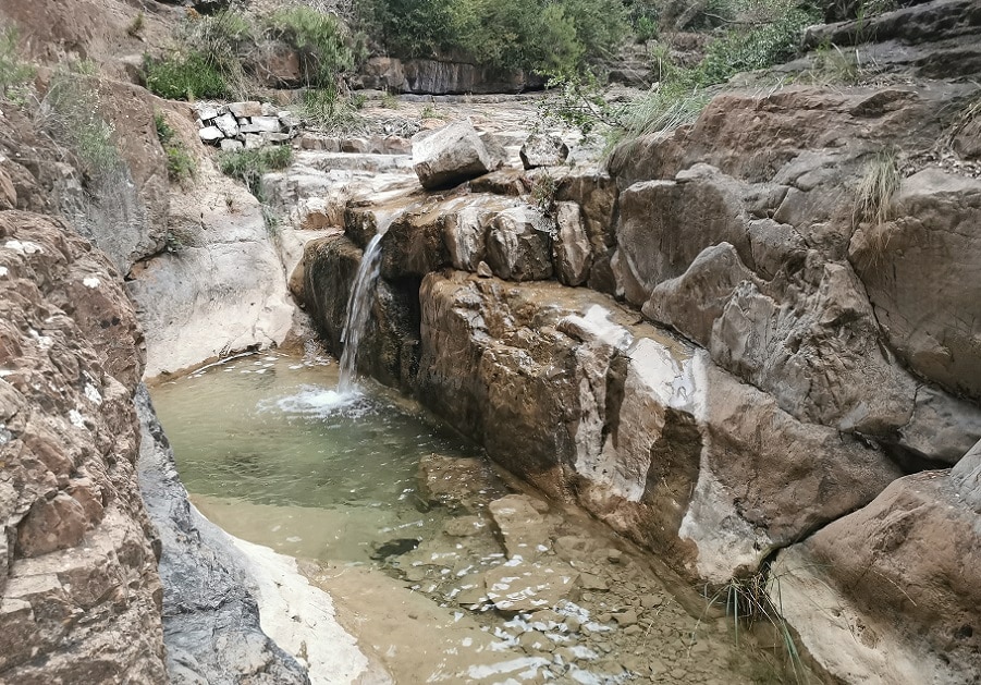 A natural rock formation is captured, featuring a small waterfall flowing into a shallow pool of clear water. Surrounding vegetation is present, enhancing the serene atmosphere. The rocky landscape reflects varied textures, with smooth and rough surfaces visible throughout.