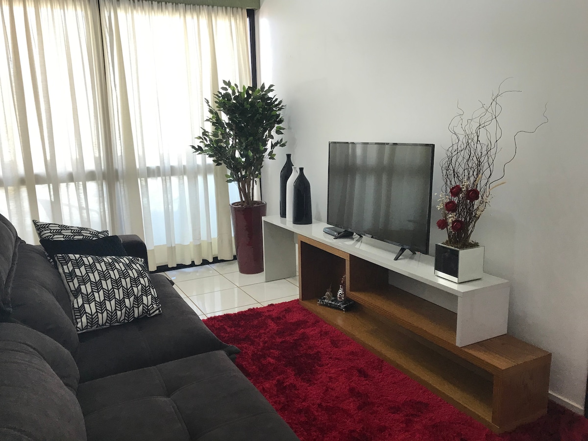 A living area is showcased with a plush gray sofa in front of a wooden TV stand. A television is positioned on the stand, complemented by decorative plants and vases. Natural light enters through floor-to-ceiling curtains, illuminating a vibrant red rug on the tiled floor.