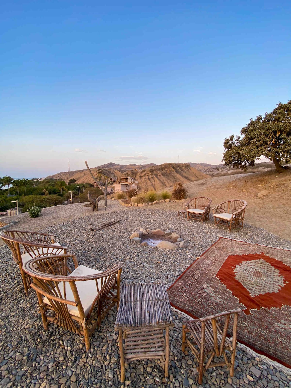 An outdoor seating area is surrounded by gravel and features four natural wood chairs positioned around a fire pit. A small table stands beside one chair, while a patterned rug adds warmth to the scene. Gentle hills and a clear sky create a serene backdrop.