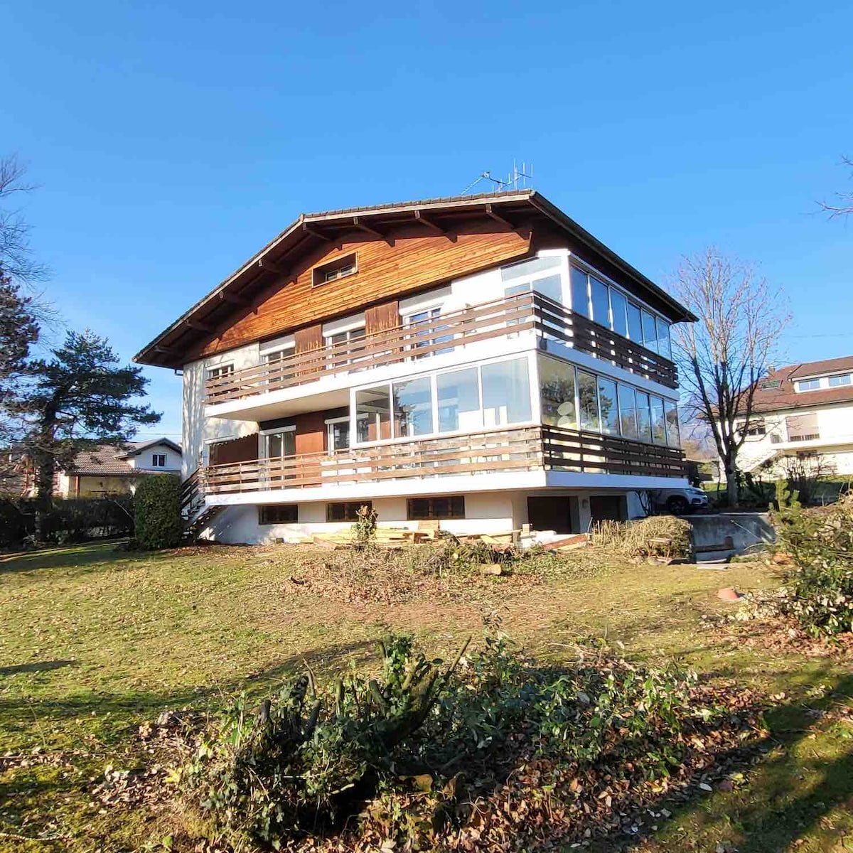 A spacious chalet is set in a green landscape, featuring multiple large windows and balconies that provide ample natural light. The building's wooden elements and modern design harmonize with the surrounding scenery, inviting guests to enjoy the outdoor space.