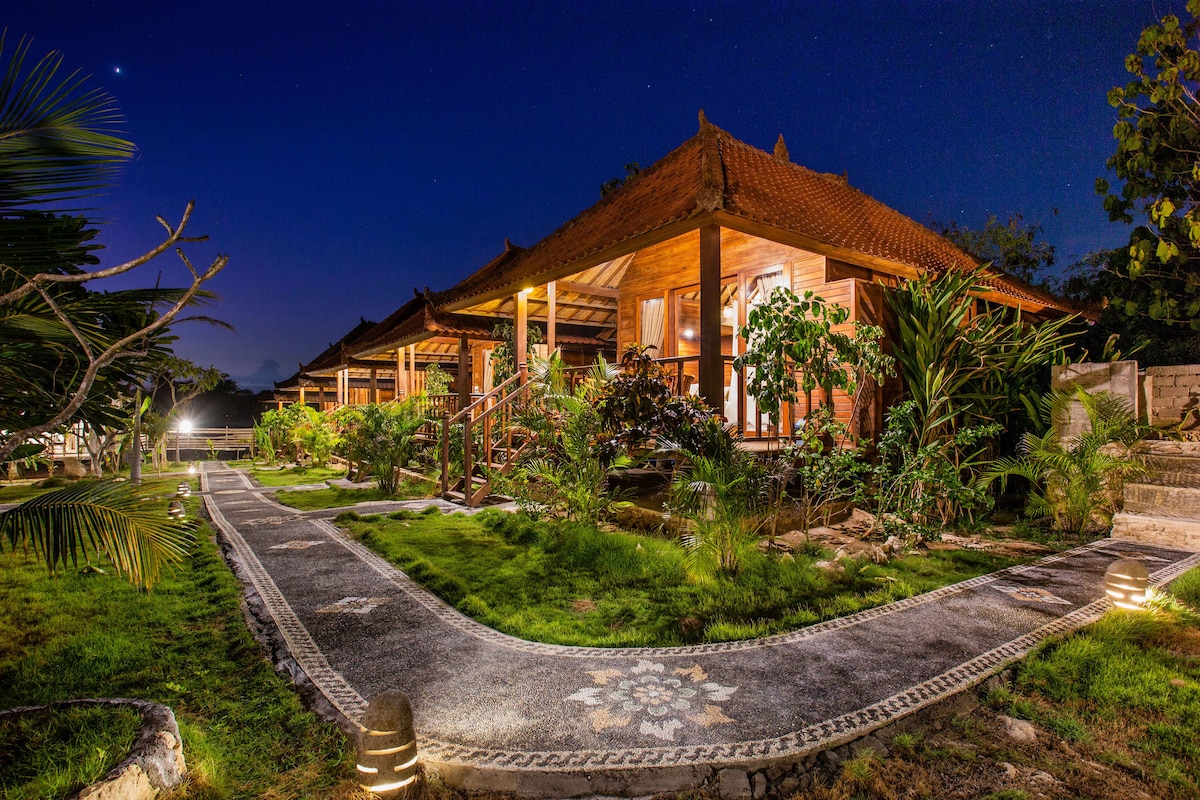 A collection of Balinese wooden cottages is nestled amidst tropical gardens, illuminated softly by outdoor lighting. A winding stone pathway guides guests toward the cottages, framed by lush greenery and small trees under a night sky. Warm light glows from the windows, inviting relaxation.