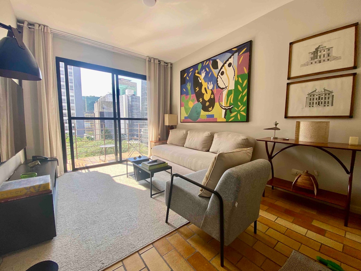 A living area is presented with a neutral sofa and accent chair, accompanied by a small coffee table. A large art piece decorates one wall, while a glass door leads to a balcony offering city views. Natural light enhances the room's warm tones.