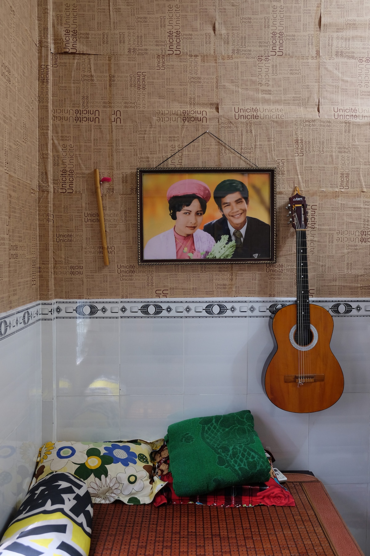 An intimate corner of a room features a neatly arranged sleeping area with vibrant patterned blankets. A wooden guitar is displayed on the wall beside a framed photograph. Soft lighting enhances the warm tones of the natural textures, creating a cozy atmosphere.