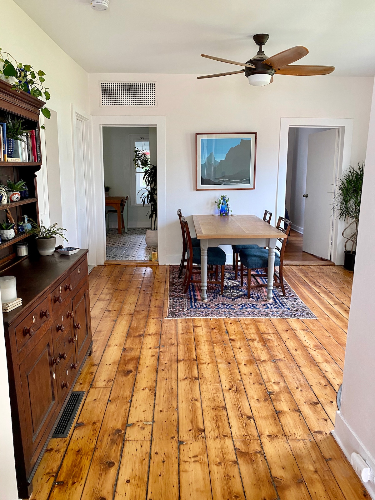 A dining area features a wooden table surrounded by six chairs on a patterned rug. A ceiling fan is present above, and a bookshelf with decorative items is positioned against a wall. Natural light is visible through doorways leading to other rooms.