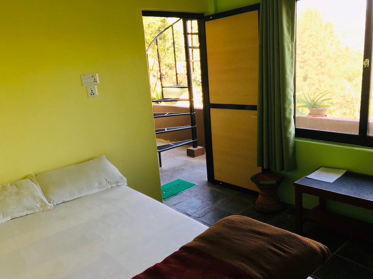 A comfortable bedroom features a spacious bed with light bedding against vibrant green walls. A double door opens to a private balcony, offering views of the surrounding nature. A small desk and chair are positioned nearby, enhancing the functional space.