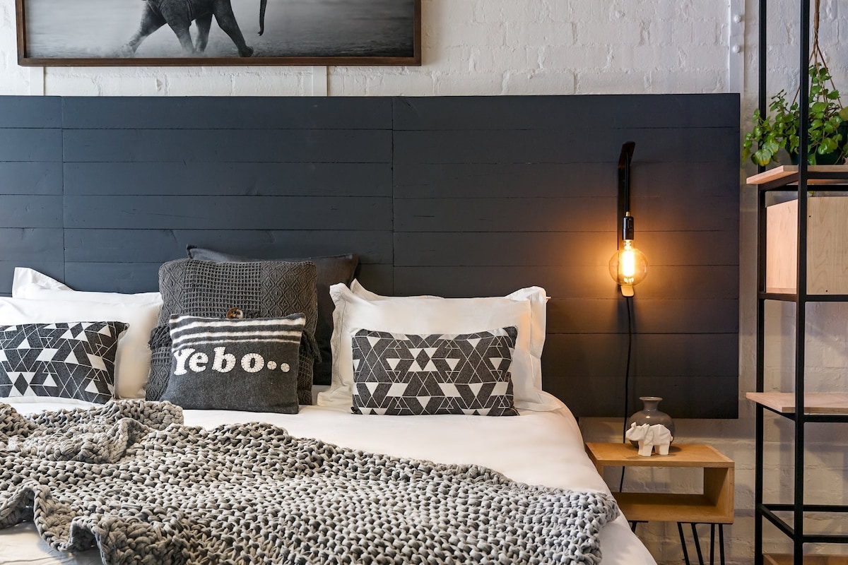 A spacious bed is adorned with a textured gray throw and patterned cushions. A bedside table with a lamp illuminates the area, while a modern shelving unit displays greenery and decorative items. A striking wall art piece featuring an elephant adds character to the room.