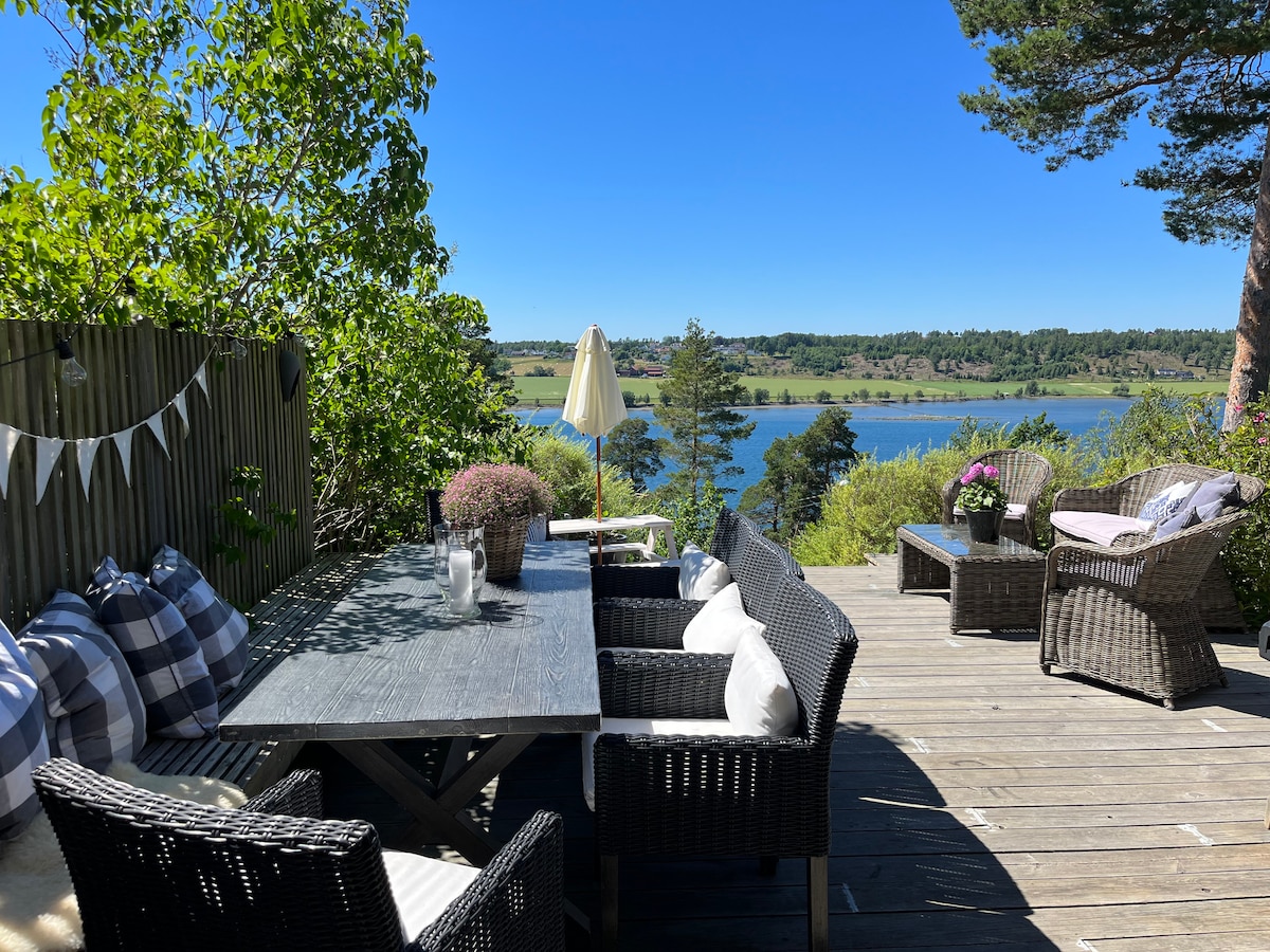 An outdoor seating area features a large wooden table surrounded by dark wicker chairs and cozy cushions. A comfortable lounge set is positioned nearby. Lush greenery frames the view of the serene water and rolling landscape under a clear blue sky.
