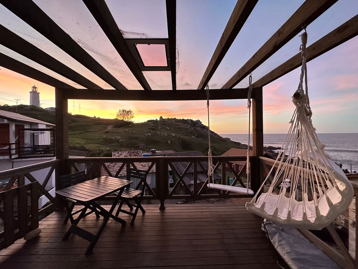 A spacious wooden deck offers a captivating view of the sunset over the ocean, with a lighthouse in the distance. A swing chair hangs beside a wooden table and chairs, creating a comfortable space for relaxation while enjoying the scenery.