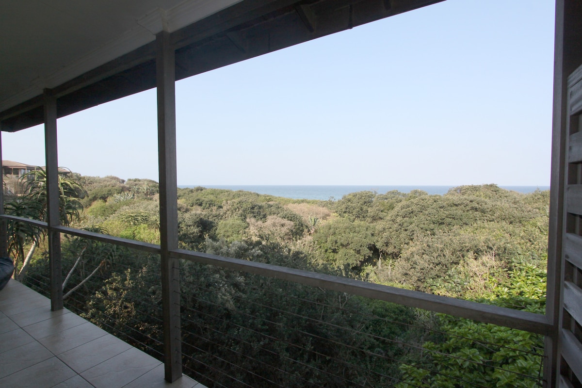 A spacious balcony provides views of lush greenery leading to the ocean. The horizon showcases a clear blue sky meeting the sea, creating a serene backdrop. The railing features a modern design, enhancing the open-air feeling of the space.