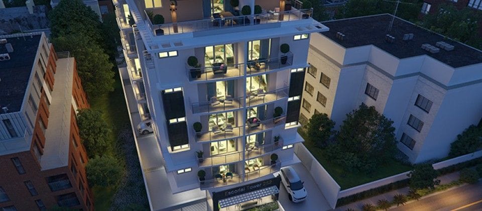 A modern building features multiple balconies with greenery, illuminated by warm lighting. The structure showcases a combination of glass and concrete, highlighting its contemporary design. A clean, organized parking area is visible at the entrance, surrounded by well-kept landscaping.