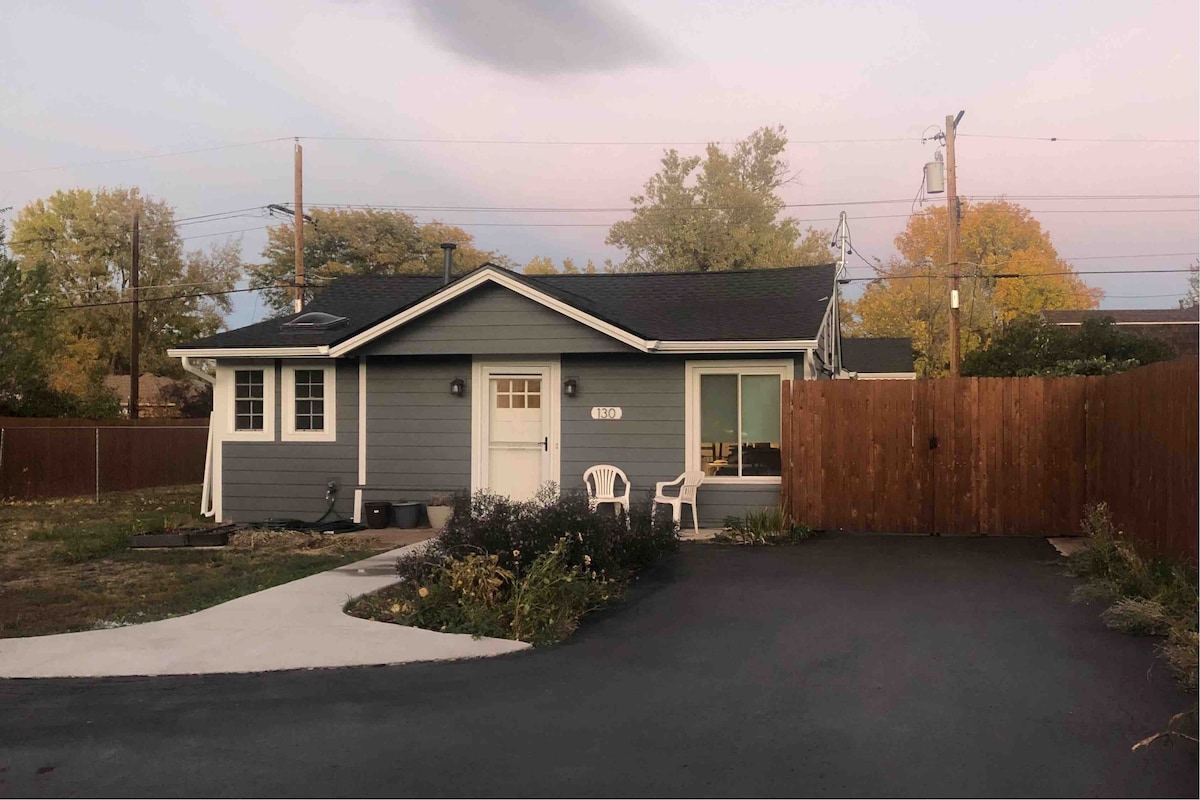 The exterior of a single-story bungalow is shown, featuring a gray facade and large windows. A small driveway leads to a spacious parking area. A wooden fence encloses a backyard, complemented by shrubs and a pair of outdoor chairs positioned on a concrete path.