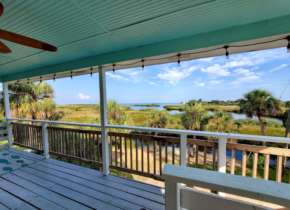 A spacious porch offers views of lush greenery and the shimmering Gulf of Mexico. Palms sway gently in the breeze, while a wide expanse of sky contributes to a serene atmosphere. Soft lighting decorates the porch railing, enhancing the connection to nature.