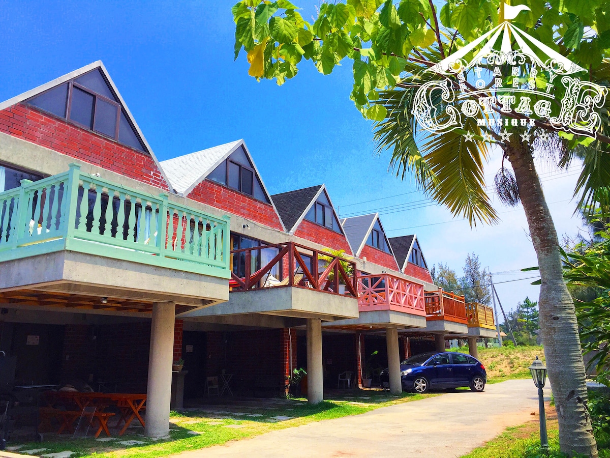 Three distinct brick cottages are visible, each featuring a balcony and unique color accents. The cottages are positioned parallel to a grassy area, with a parked car in the foreground. Clear blue skies and greenery create a relaxed outdoor environment.