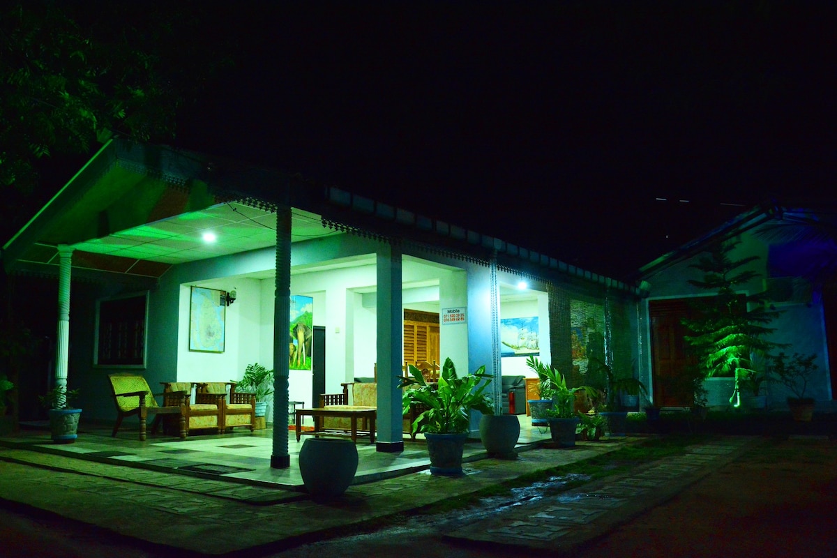 The exterior of the property is showcased at night, illuminated by green lighting. A spacious porch is visible, featuring wooden seating and decorative potted plants. The entrance is framed by a covered area, leading towards a well-maintained pathway and garden.