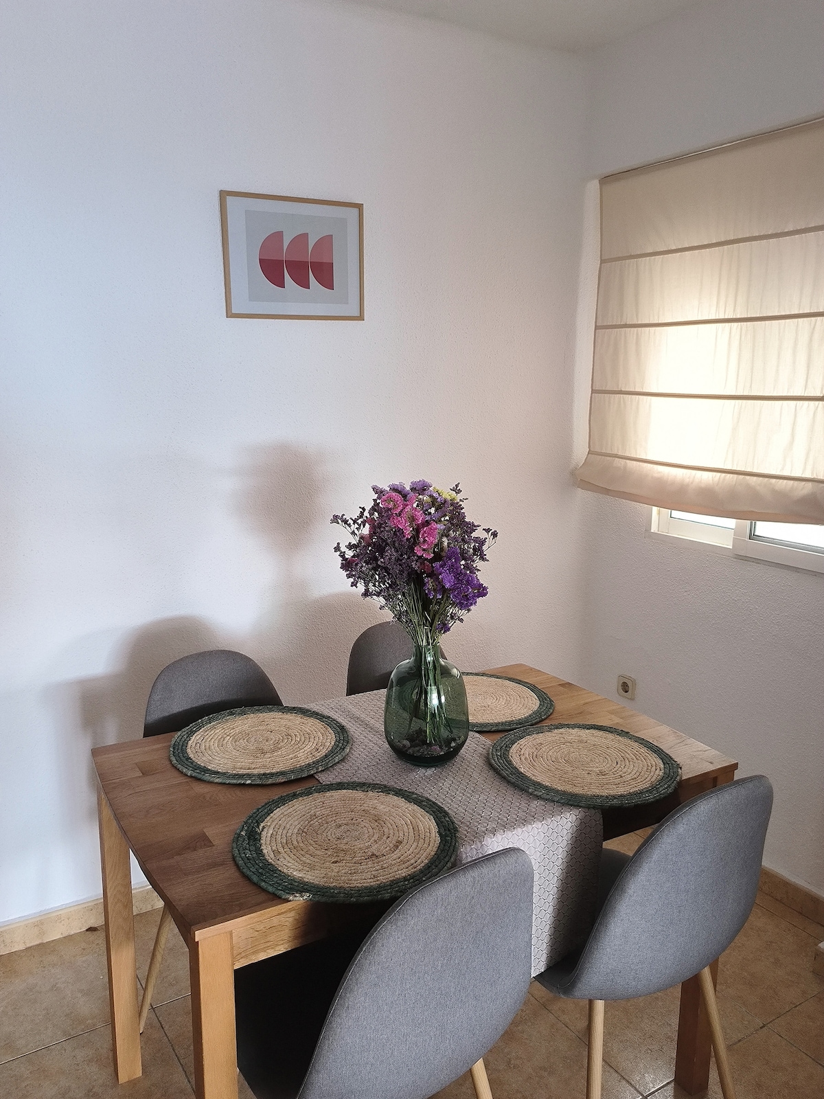 A dining area is presented with a wooden table surrounded by four grey chairs. A round centerpiece features a vase filled with colorful flowers. Place settings with woven placemats and plates are arranged on the table, illuminated by natural light from a nearby window.