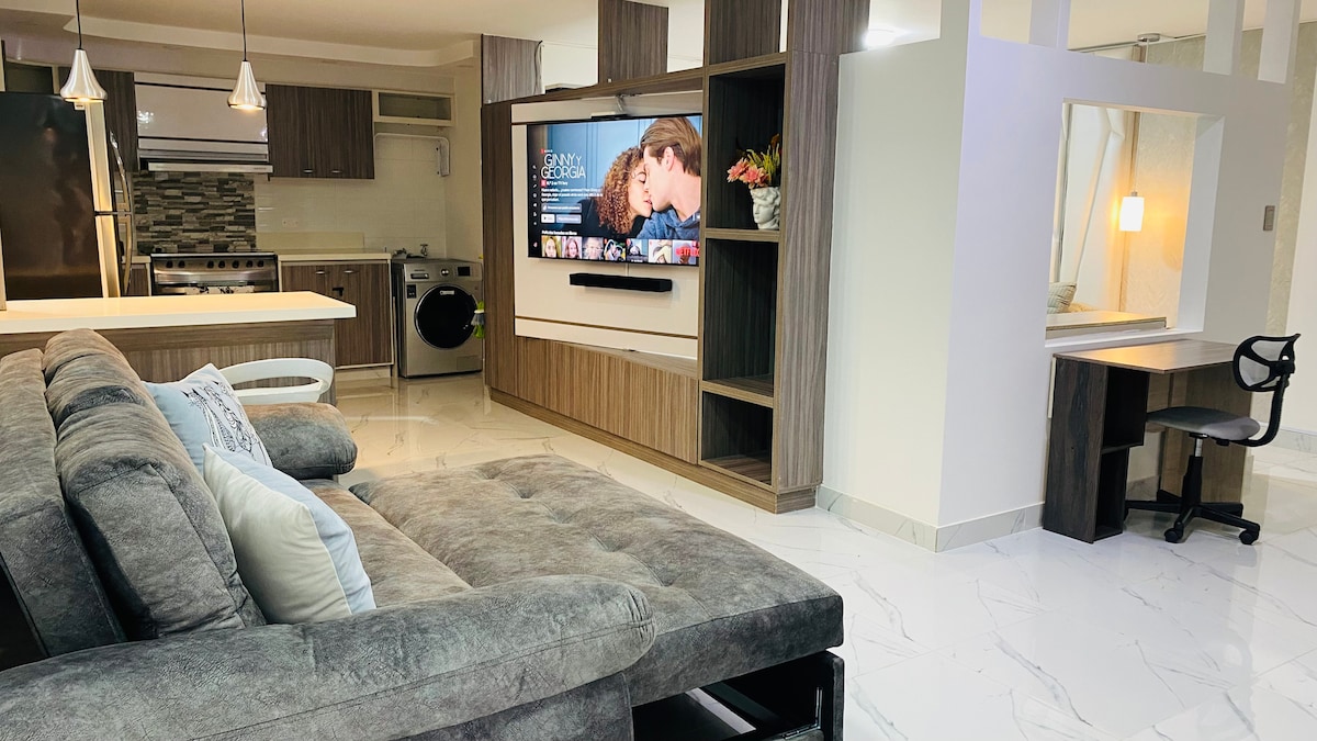 A modern open-plan living area features a comfortable gray sofa positioned towards a large mounted television. Nearby, a work desk with a chair and a fully equipped kitchen with sleek cabinetry are visible. Natural light illuminates the space through strategically placed lighting fixtures.