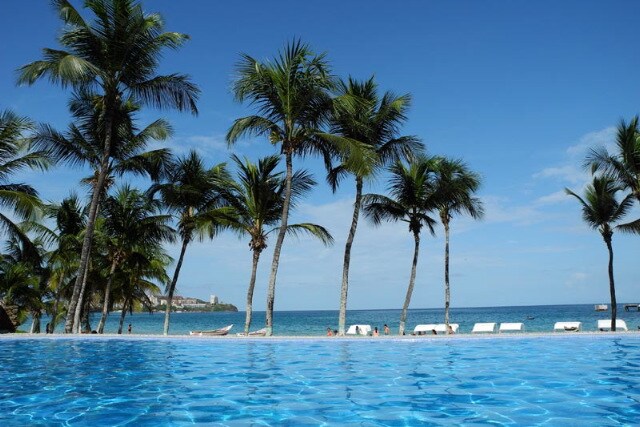 The infinity pool features clear blue water, reflecting the sky above. Palm trees surround the area, swaying gently in the breeze. The ocean is visible in the background, with distant boats and white sandy shores adding to the serene atmosphere.