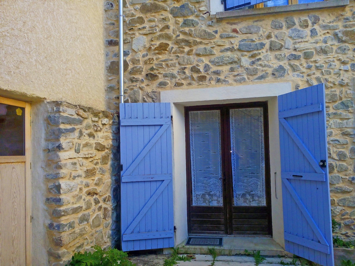 A traditional stone exterior features a pair of open blue shutters framing a wooden door with glass panels. The entrance is highlighted by a small welcome mat on a stone pathway, surrounded by lush greenery.