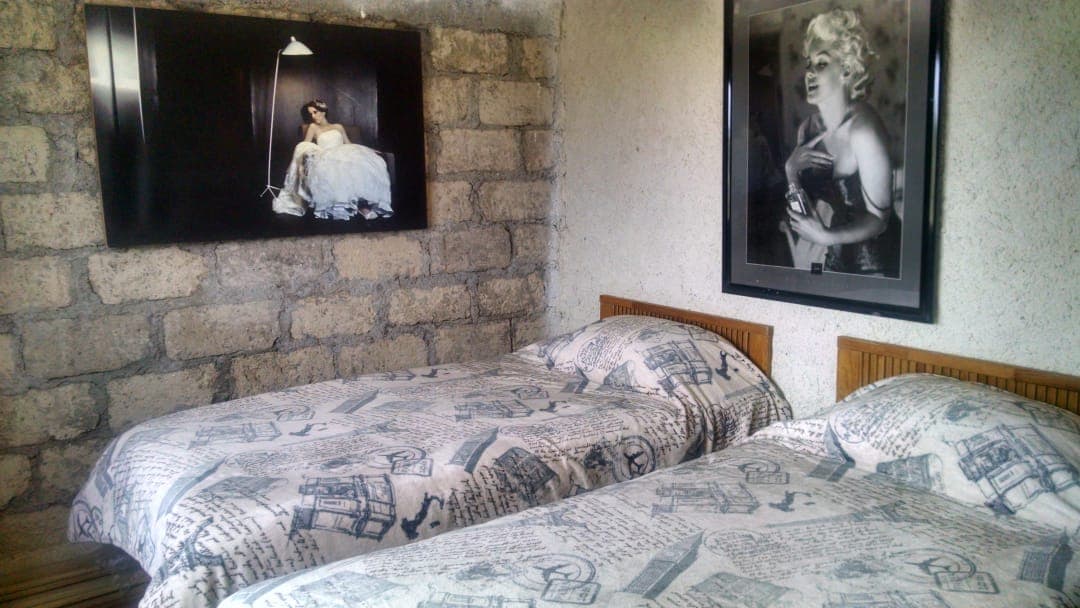 A bedroom features two neatly made beds, each adorned with patterned bedding. Black and white photographs are displayed on the walls, enhancing the room's aesthetic. Textured stone walls contribute to a rustic feel, complemented by soft lighting from a hanging lamp.