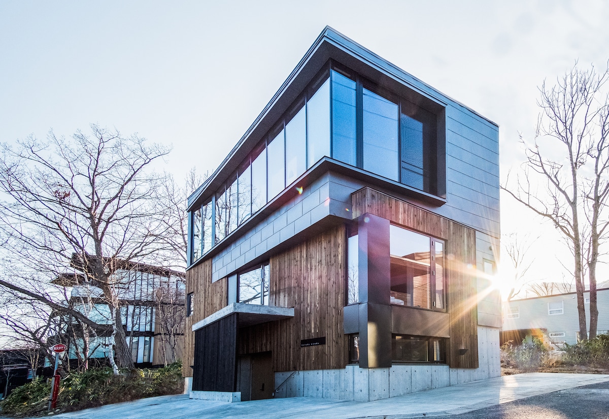 The exterior of the modern three-story home features a combination of sleek metal and natural wood materials. Large glass windows provide ample natural light, while a driveway and landscaping enhance the welcoming atmosphere. The structure captures the sun's rays, adding warmth to the overall design.