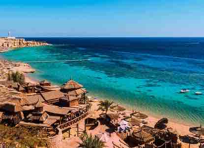 A scenic view of a beach featuring soft sand and clear blue waters. Comfortable lounge areas with thatched roofs are arranged along the shore, surrounded by palm trees. The horizon showcases a vibrant turquoise sea, blending seamlessly with the bright sky.