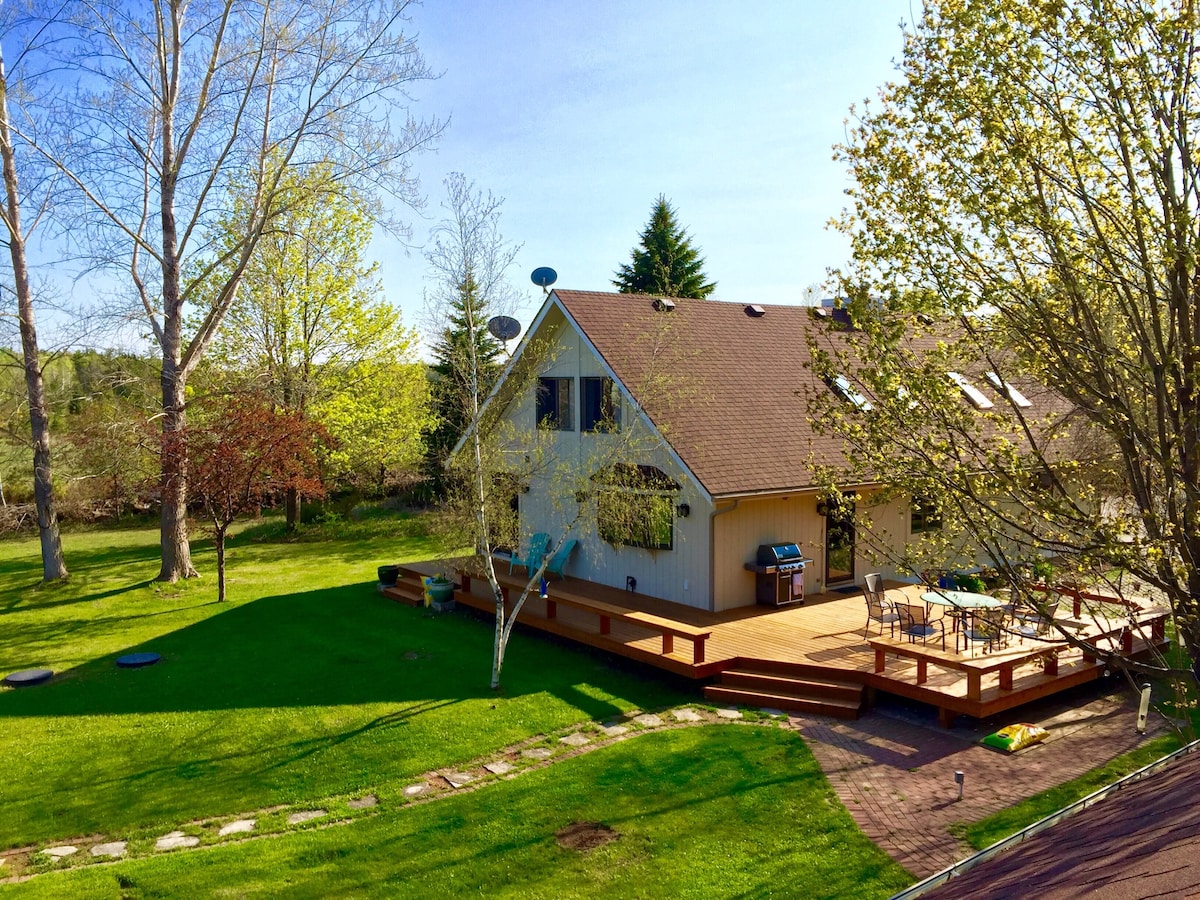 A well-maintained yard features a spacious wooden deck and patio area, equipped with outdoor seating and a grill. Lush green grass surrounds the house, framed by mature trees that provide shade. The property is bathed in natural light under a clear blue sky.