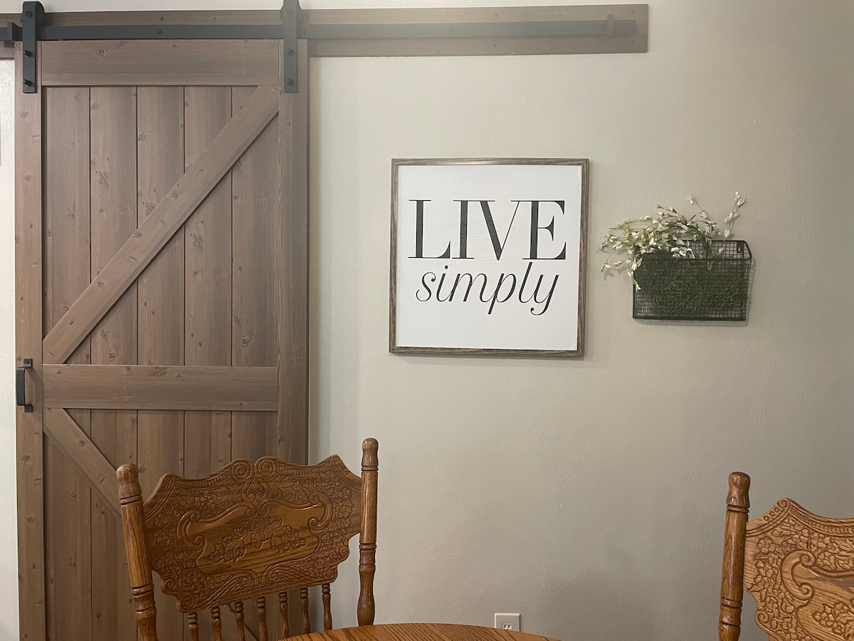 A rustic wooden door with a sliding mechanism is positioned next to a framed sign that reads 'LIVE simply.' A wall-mounted planter holds a small arrangement of greenery, complementing the warm wood tones of the dining chairs and table in the scene.
