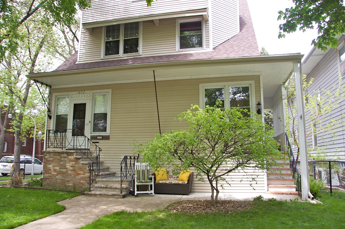 The entrance to the vintage home is framed by lush greenery, showcasing a well-maintained front yard. A charming porch features seating near the steps, inviting relaxation. Large windows provide plenty of natural light, enhancing the home's welcoming appearance.