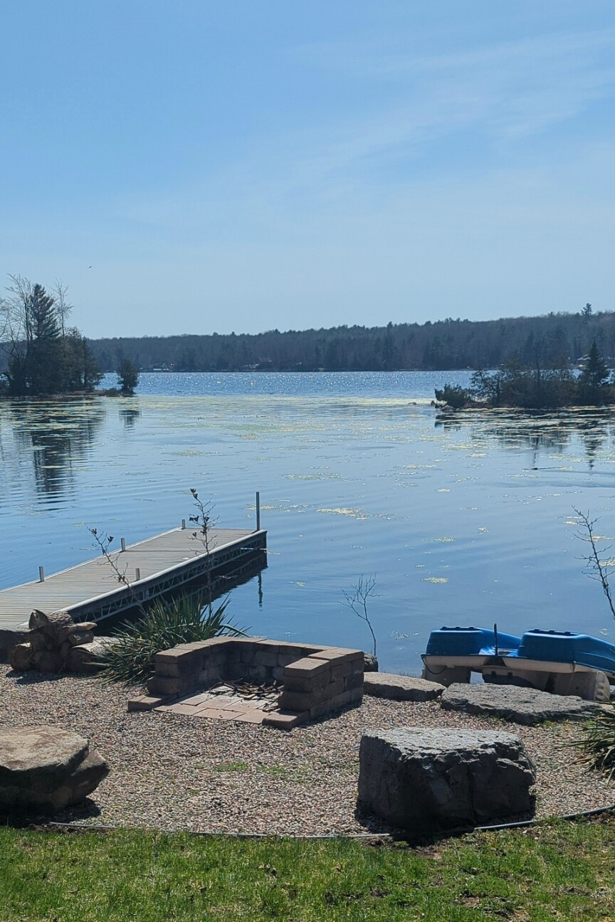 A serene lake view features calm waters reflecting the sky, surrounded by lush greenery and distant trees. A wooden dock and two blue kayaks are positioned at the water's edge. A stone firepit and landscaped garden area can be seen in the foreground.