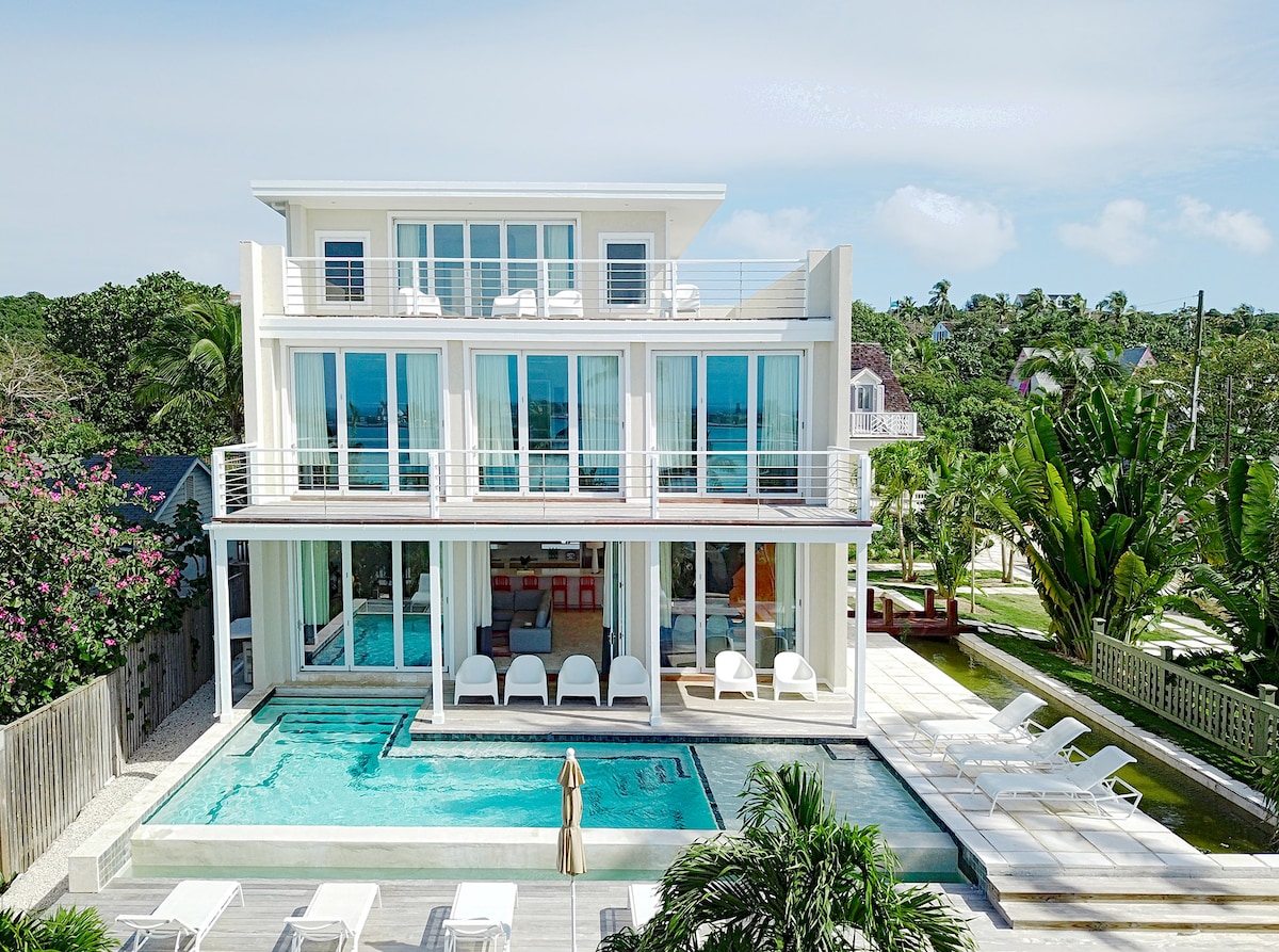A modern three-story villa is shown, featuring large glass windows and multiple balconies. An infinity pool is visible in the foreground, surrounded by lounge chairs. Lush greenery and tropical landscaping enhance the outdoor space, contributing to a serene atmosphere.