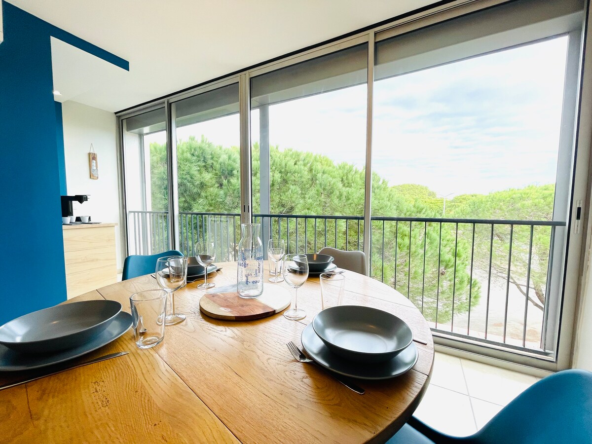 A round wooden dining table is set with plates and glasses, positioned near large glass sliding doors. Outside, views of greenery are visible through the balustrade, providing a tranquil atmosphere. Soft natural light illuminates the space, enhancing the welcoming environment of the dining area.