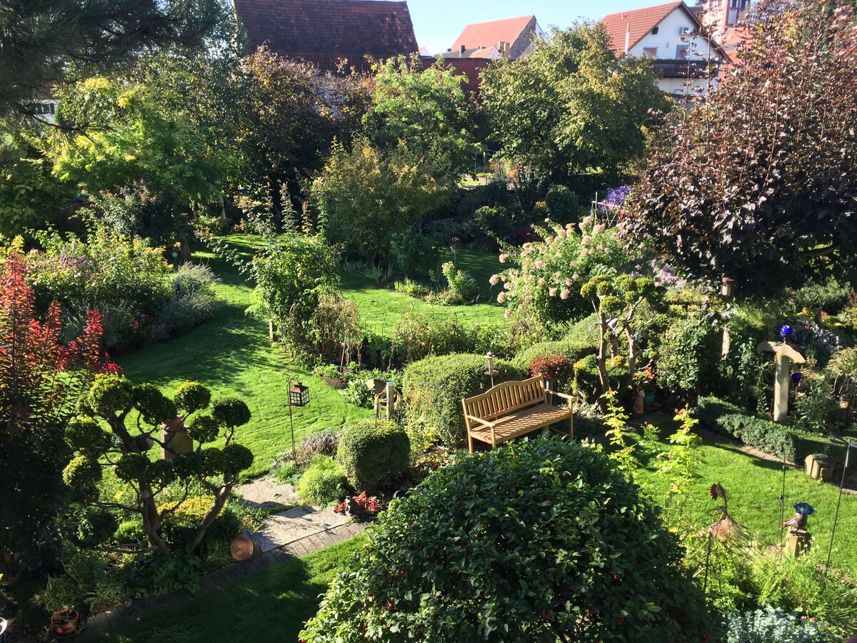 A lush garden with diverse greenery and blooming flowers creates an inviting outdoor space. A wooden bench is positioned amid the meticulously arranged plants, providing a serene spot to relax. Various ornamental features and pathways enhance the natural beauty of the 2200 sqm area.